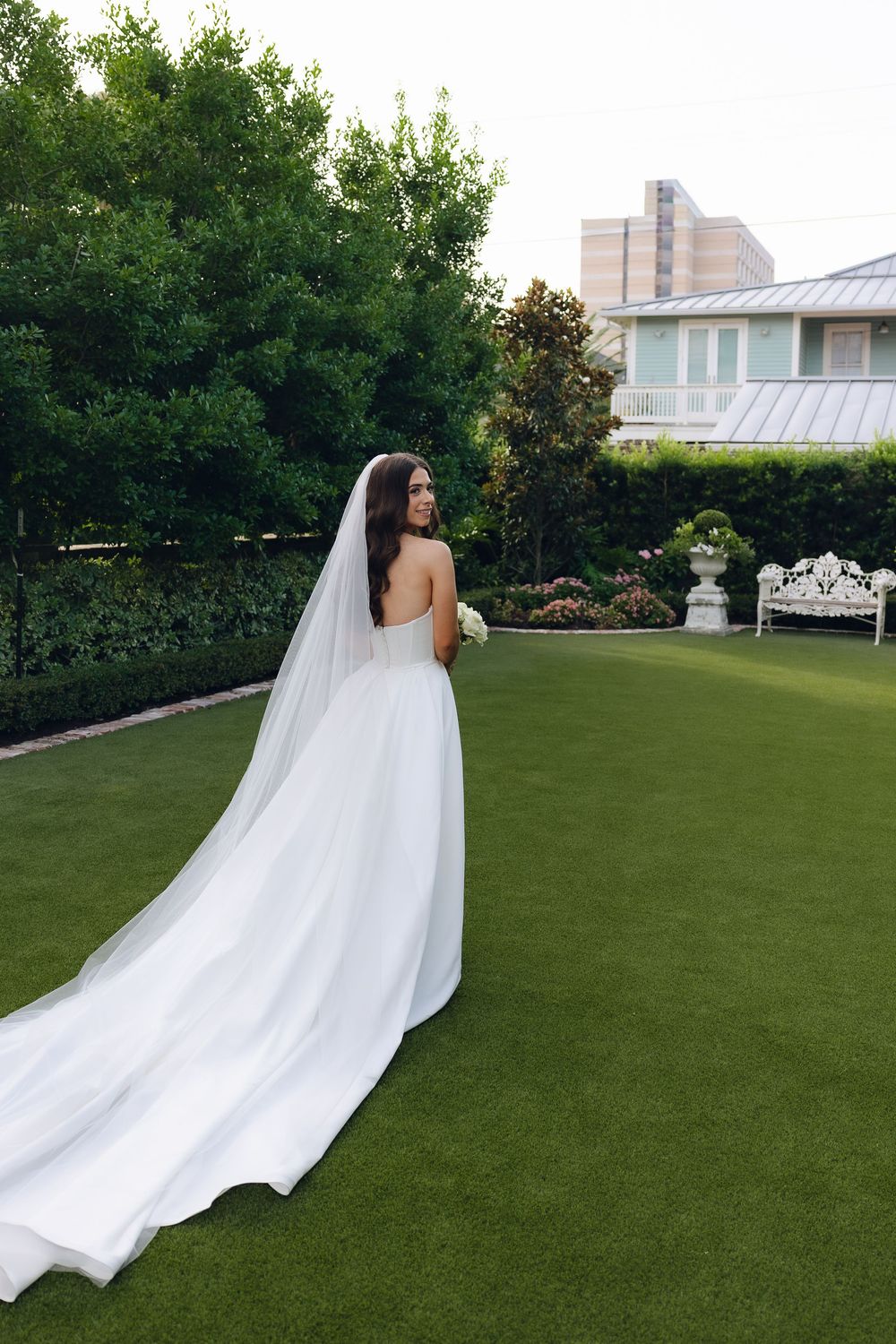 A bride in a flowing white wedding gown with a long cathedral veil walks across a manicured green lawn.