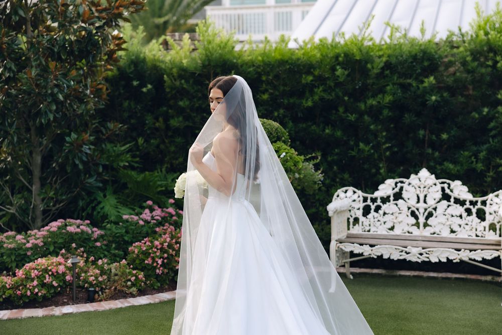 A bride in a flowing white wedding dress and long veil stands in a lush garden setting surrounded by greenery and flowers.