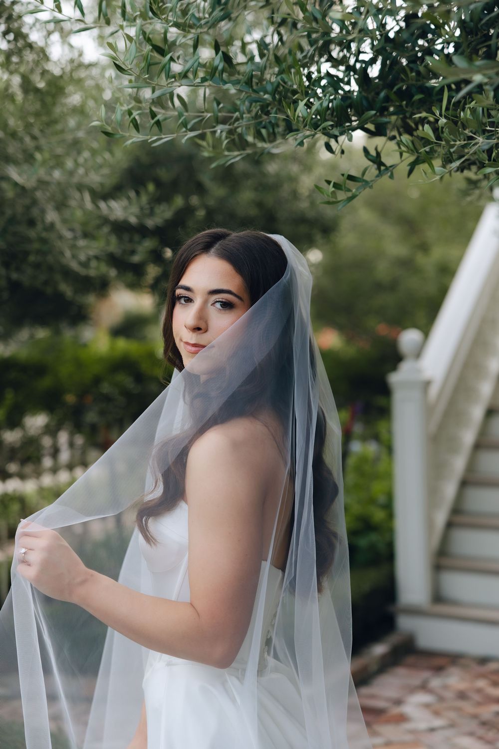 A bride wearing a flowing white veil poses in an elegant garden setting with greenery in the background.