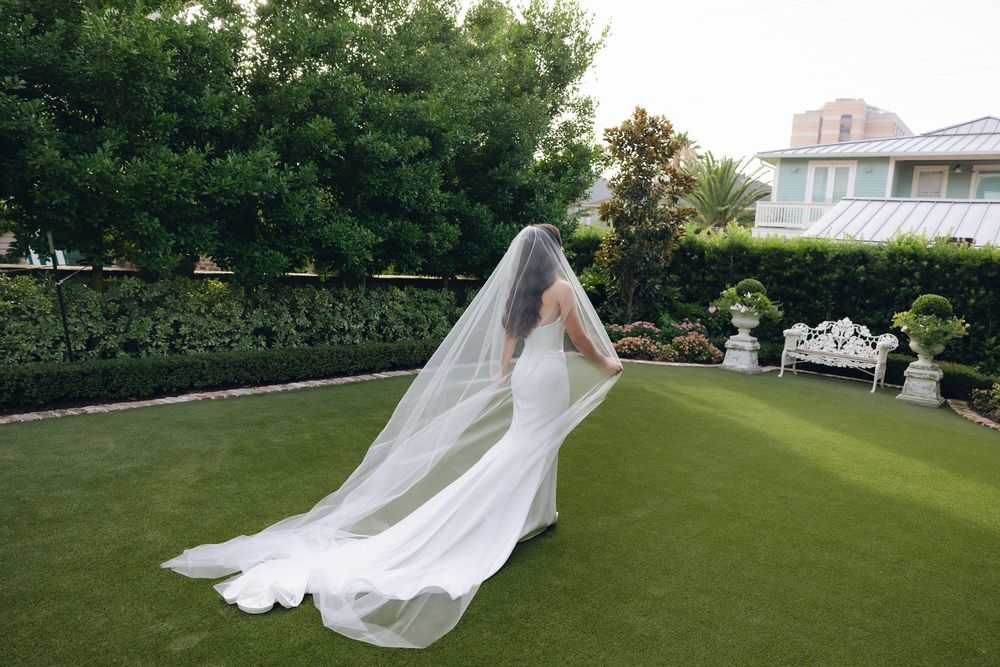 A flowing white wedding veil billows dramatically in the wind against a lush green lawn background.