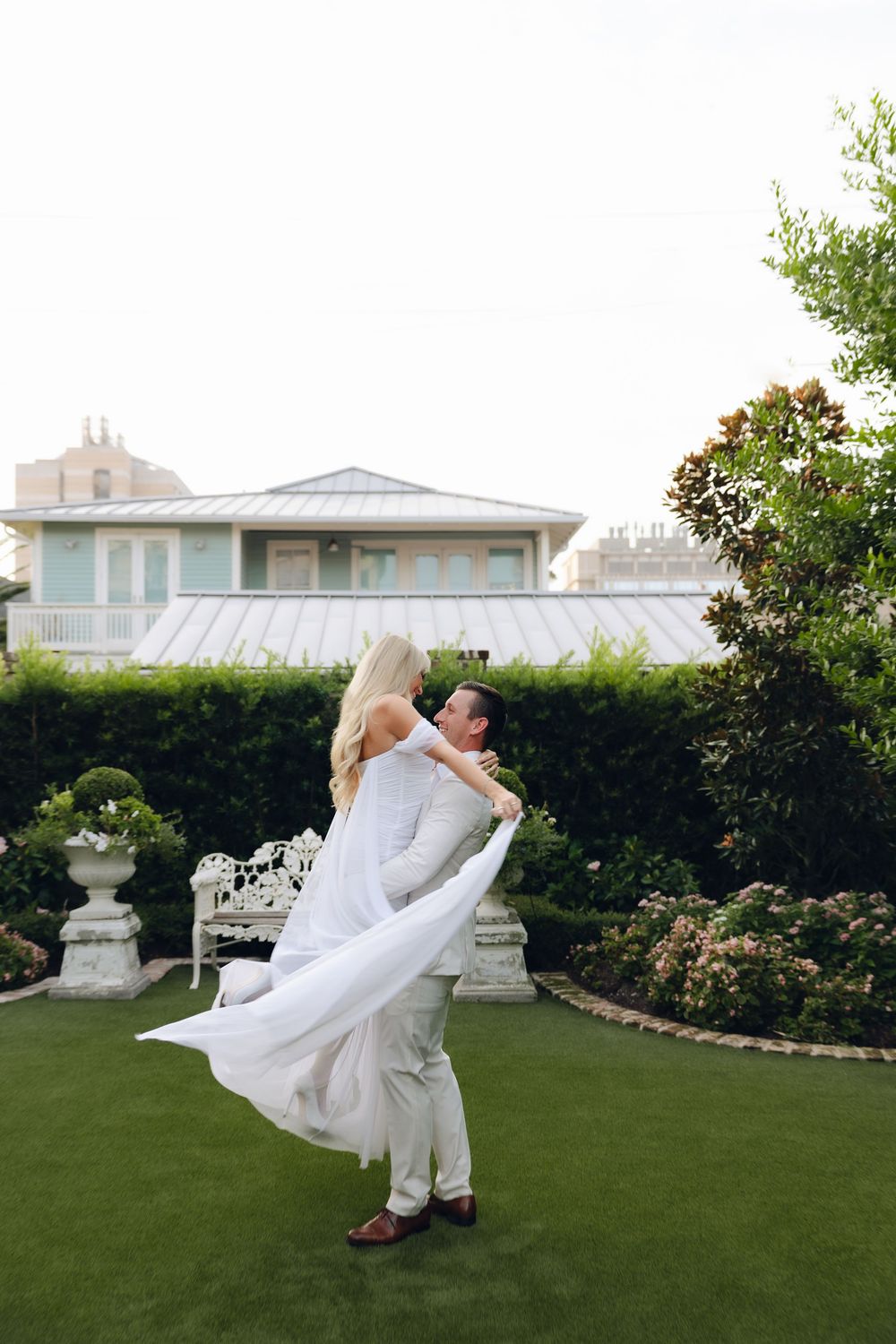 A couple in wedding attire dances and spins gracefully on a manicured green lawn surrounded by hedges and architecture.