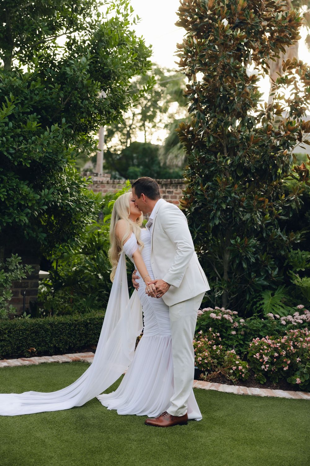 A romantic wedding couple embraces on a manicured lawn with lush greenery and flowering bushes in the background.