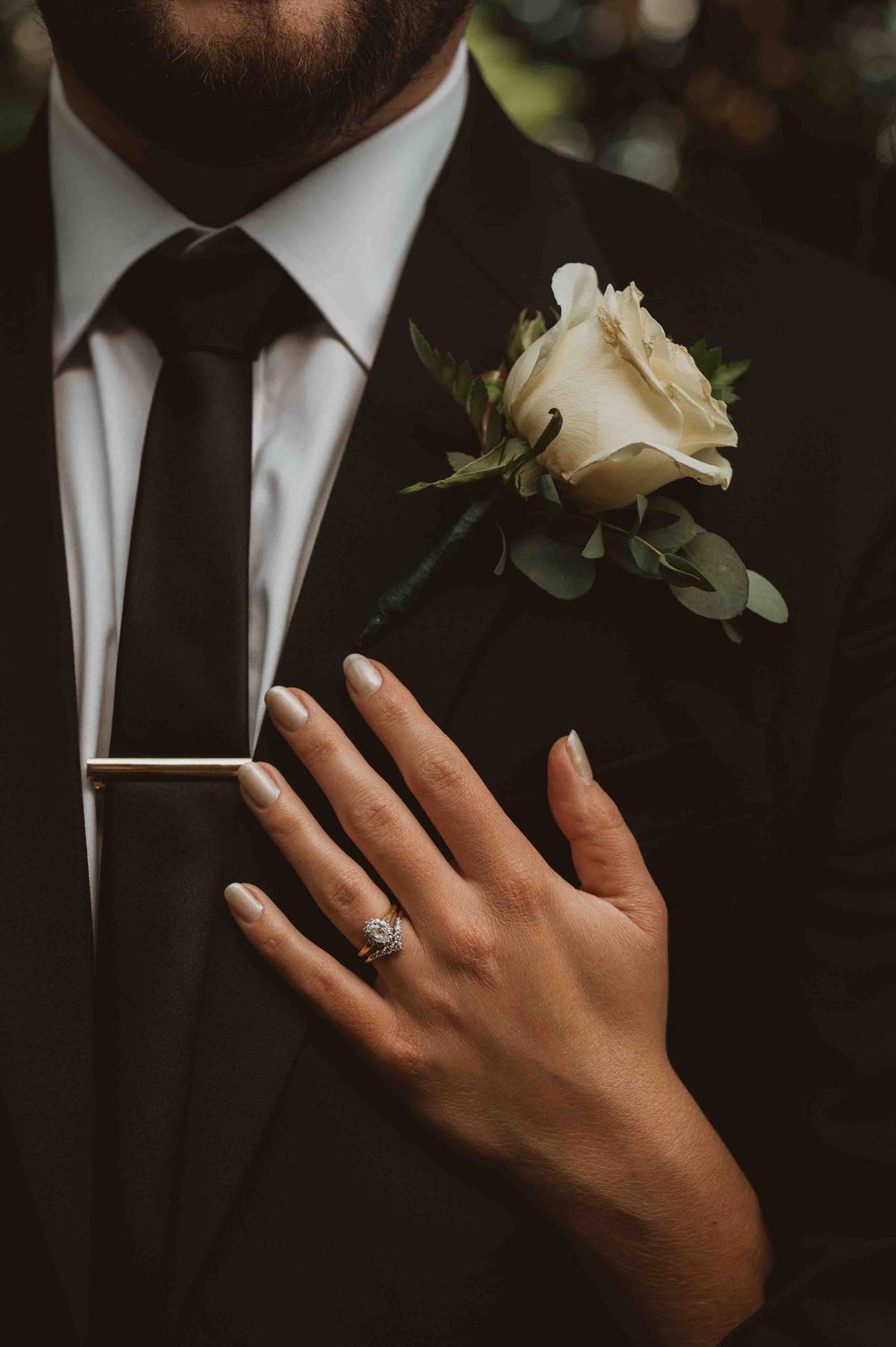 Close-up details of groom's boutonniere and suit with bride's hand showing engagement ring.