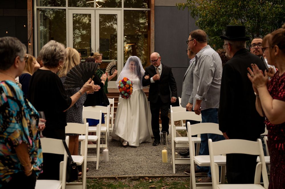 Wedding guests gather outdoors at a rustic venue with white chairs and natural decor during a ceremony.