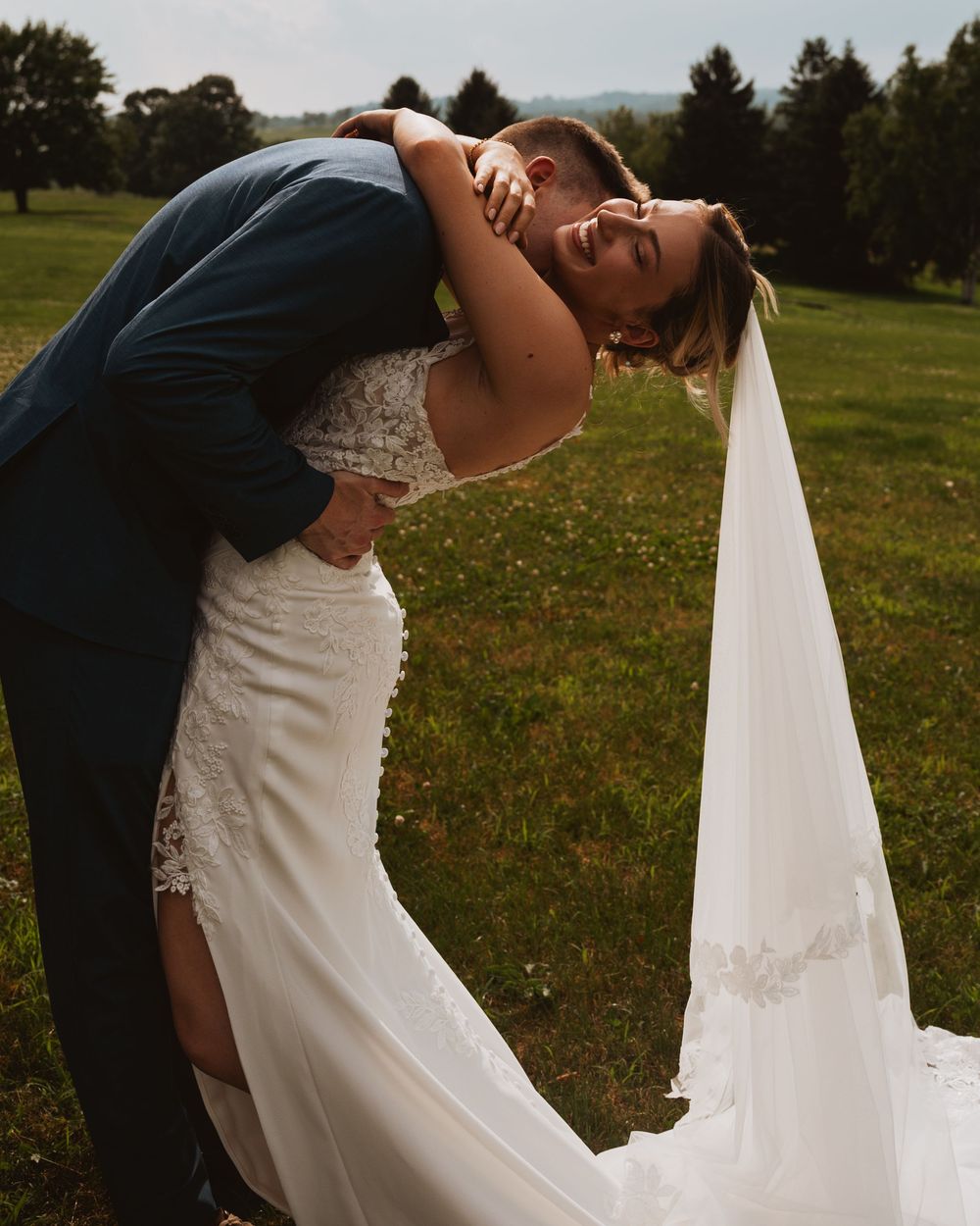 Bride and groom share a romantic dip kiss outdoors on green grass, bride in lace wedding gown with flowing veil.