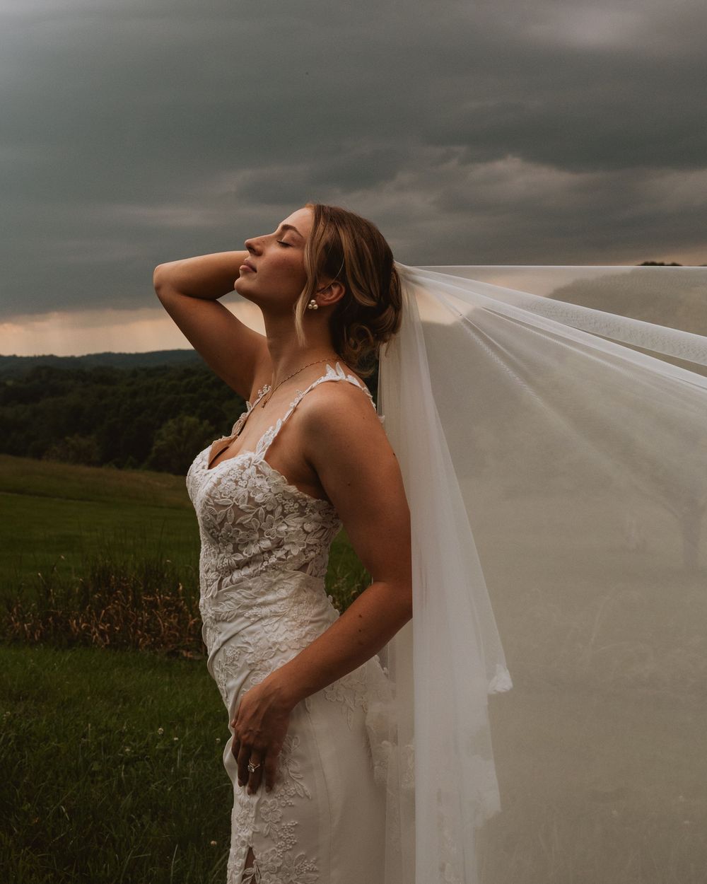 Bride in lace wedding gown and flowing veil poses dramatically against stormy sky in rural countryside setting.