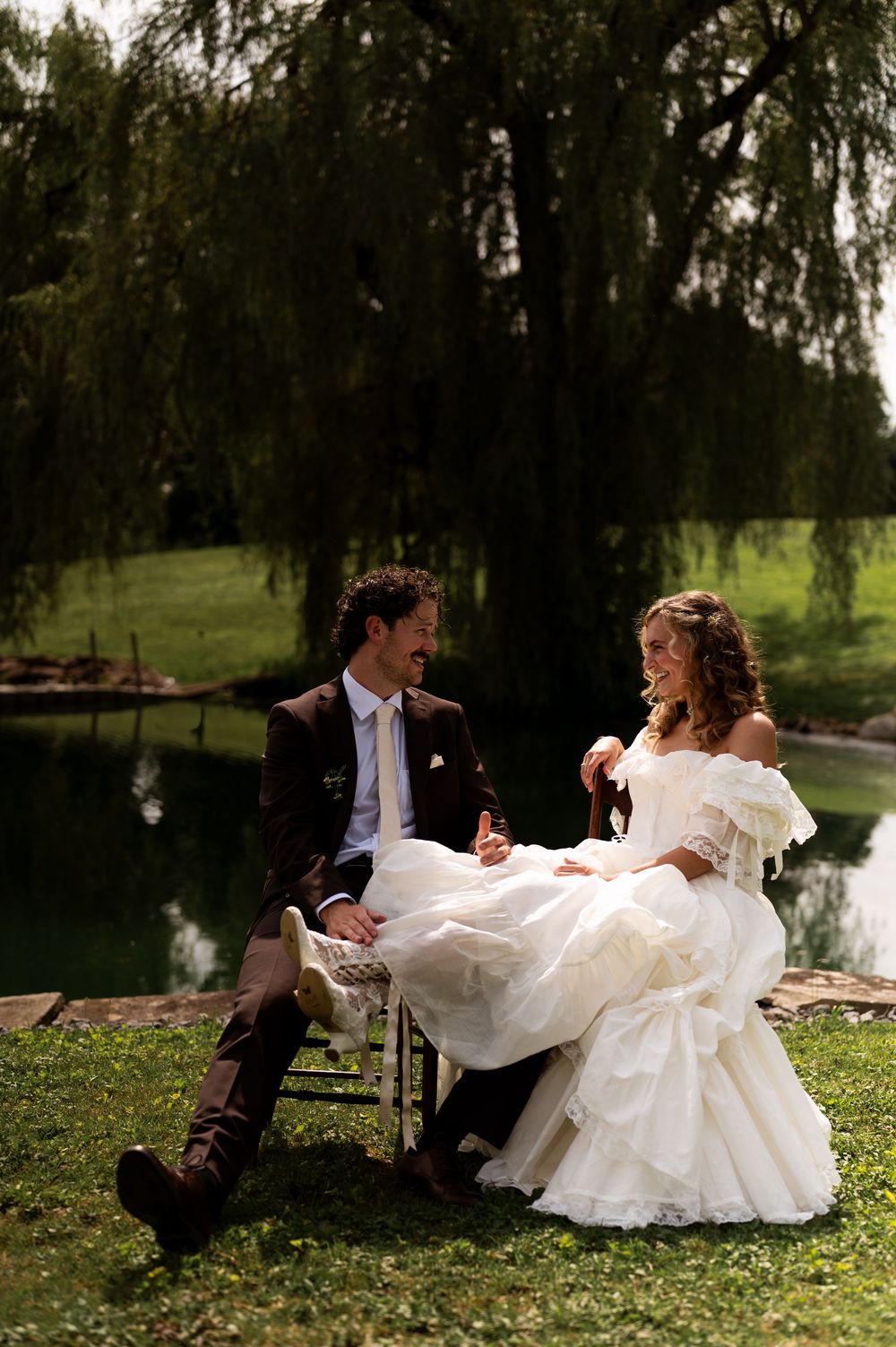Bride and groom sitting by a serene pond under a weeping willow tree, sharing a romantic moment on their wedding day.