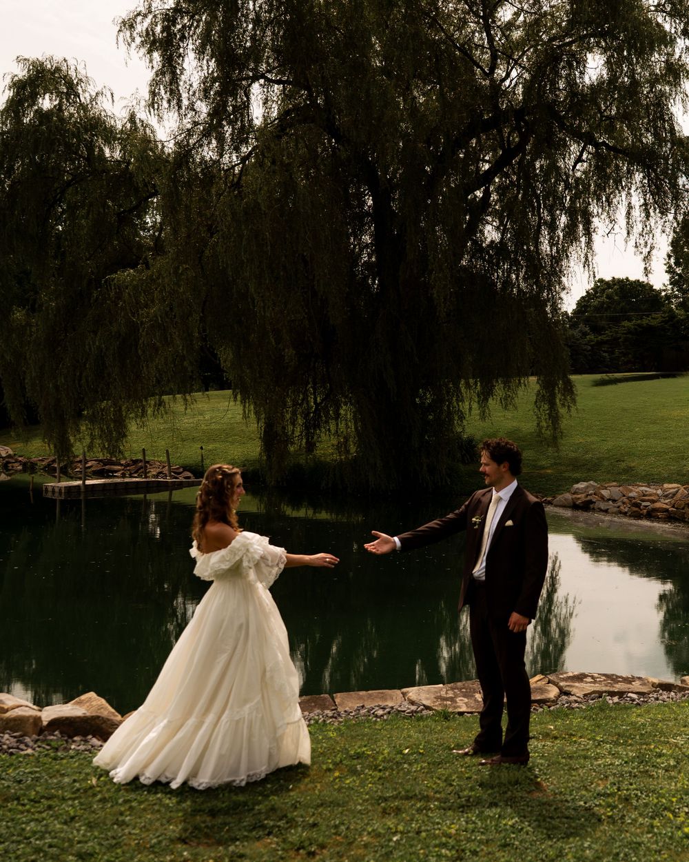 A bride and groom hold hands near a serene pond under a weeping willow tree during their outdoor wedding ceremony.