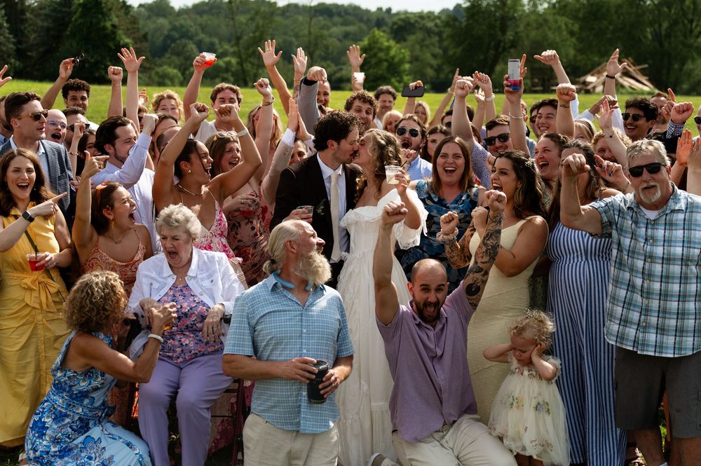 Large wedding party celebrating outdoors, guests cheering with raised arms on sunny day.