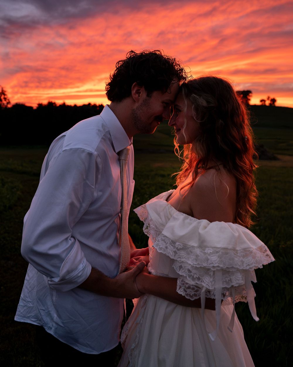 Couple sharing a romantic moment at sunset with vibrant red and orange sky over a lush green field.