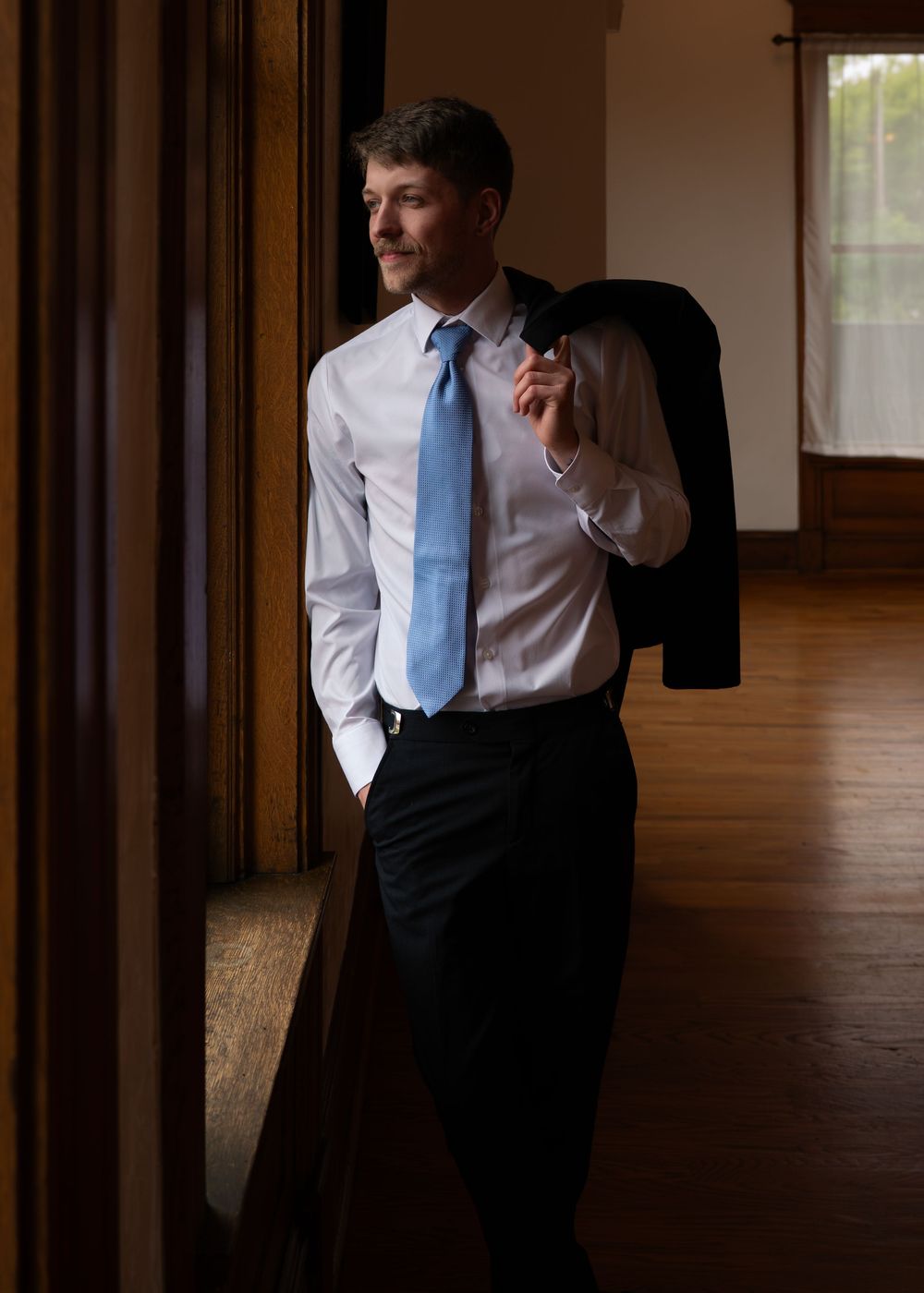Man in white shirt and blue tie holds suit jacket over shoulder, standing by window in elegant wood-paneled room.