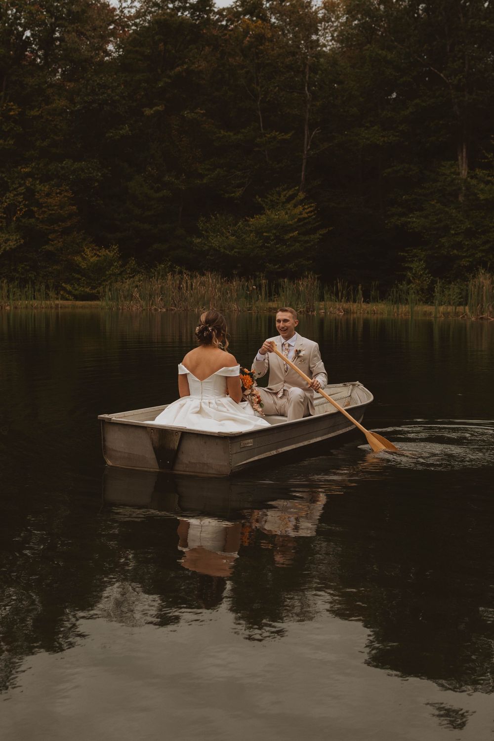 Two people relax together in a small rowboat on a tranquil lake surrounded by trees at dusk.