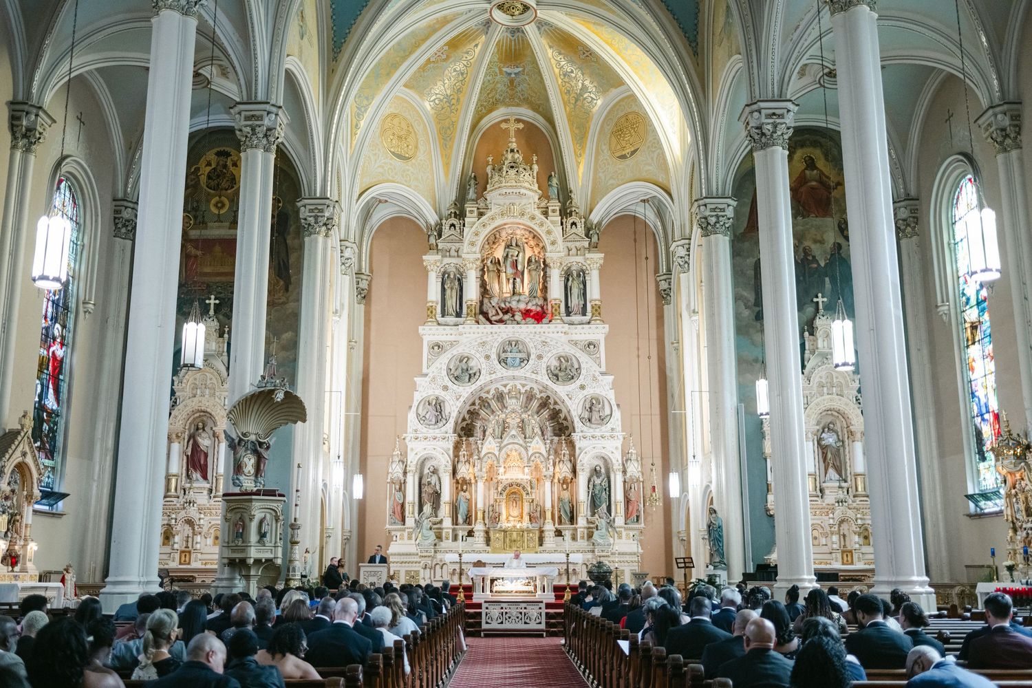 Interior panoramic view of a grand Catholic church with ornate architecture, stained glass windows, and filled pews.