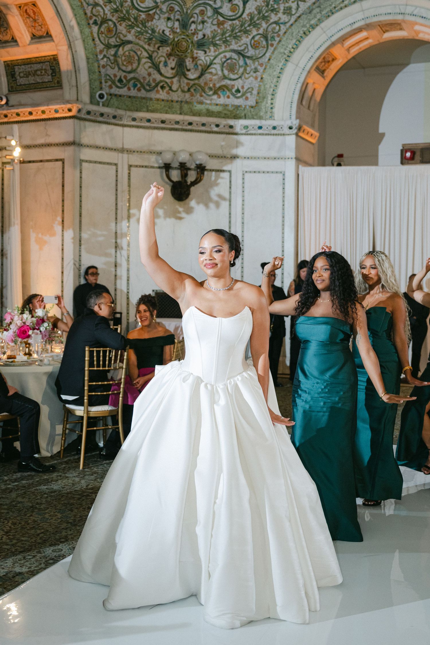 Wedding party members in teal bridesmaid dresses dance and celebrate with the bride in a white ballgown at an ornate venue.