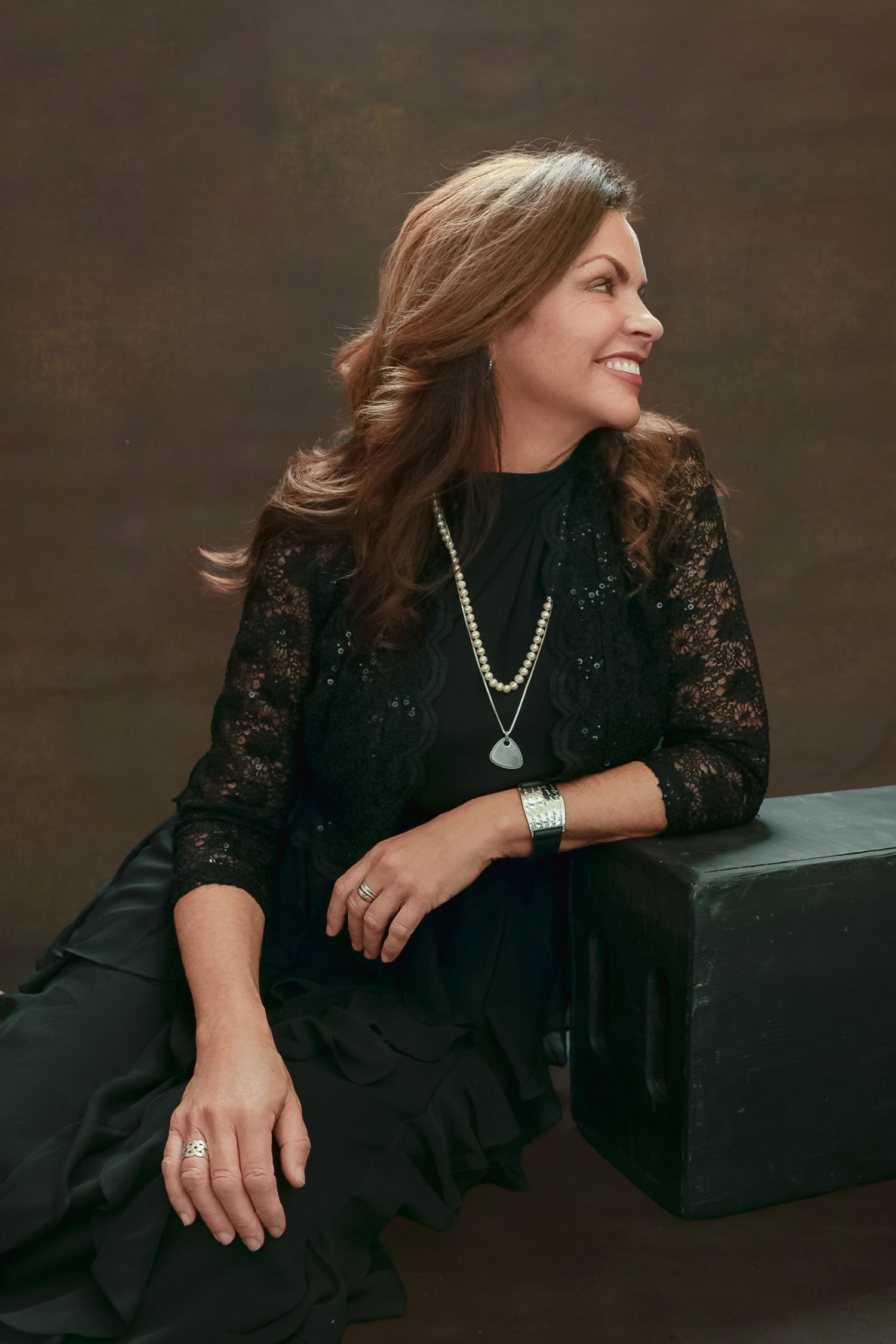 A person wearing a black lace dress and jewelry sits casually against a dark background in a series of candid poses.