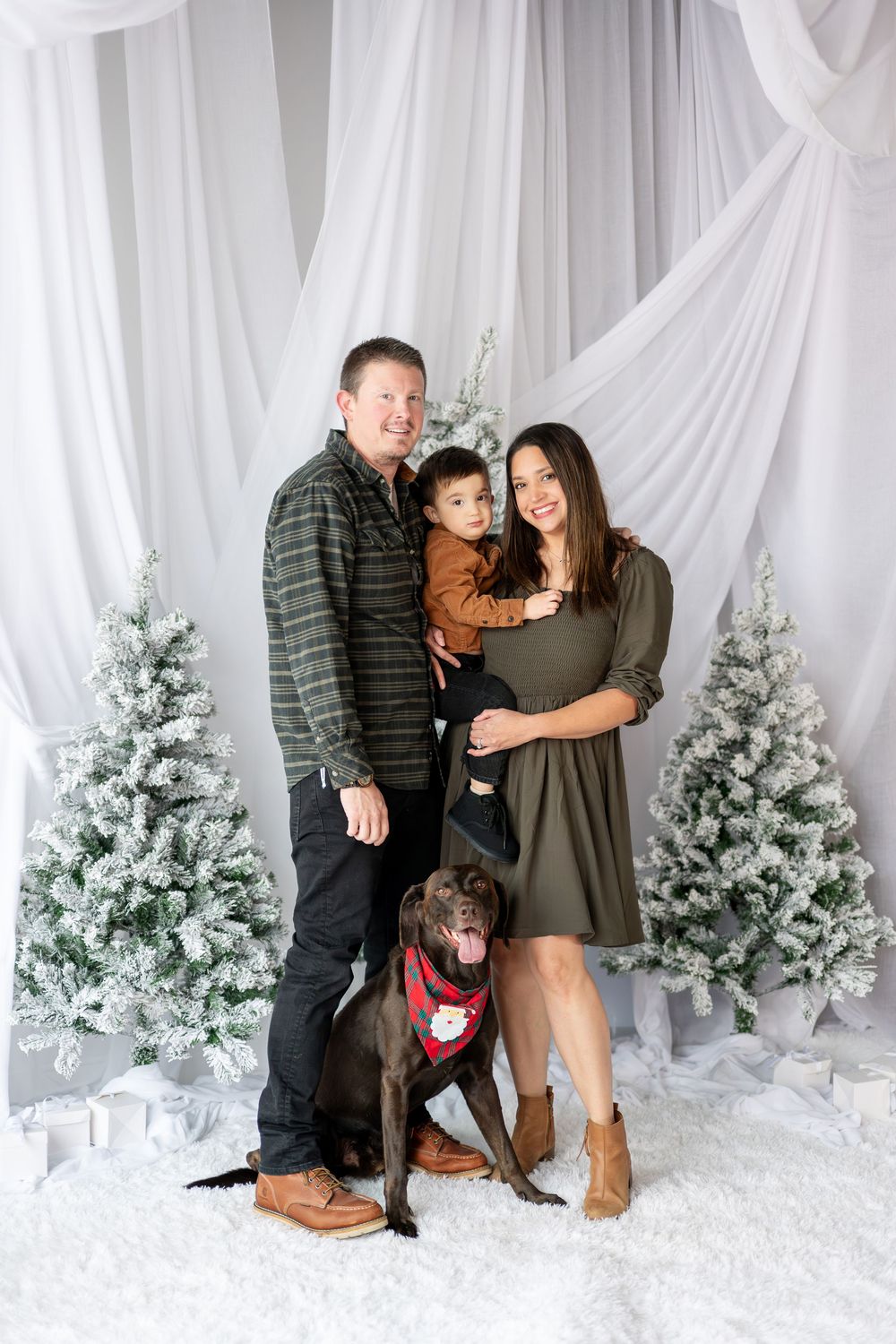 A family of three with a Labrador in a red bandana poses before white-draped snowy Christmas trees.