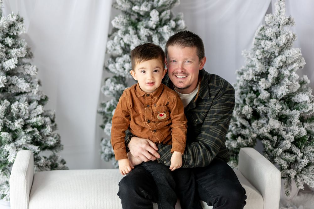 Father and young son sitting together on white bench with snow-covered Christmas trees in holiday photo studio.
