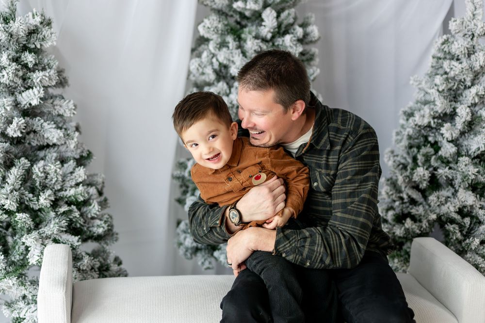 Laughing father playfully hugging his young toddler son on white bench with flocked Christmas trees behind them.