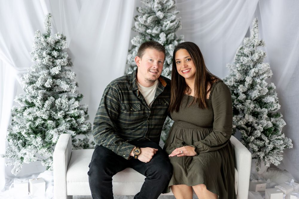 Expecting couple sitting together on white bench with snow-flocked Christmas trees in holiday maternity photo session.