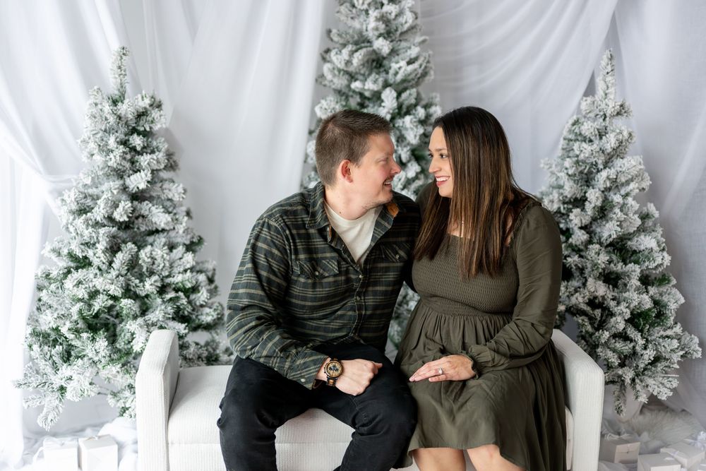 Smiling expecting couple gazing at each other on bench surrounded by snow-flocked Christmas trees in studio.