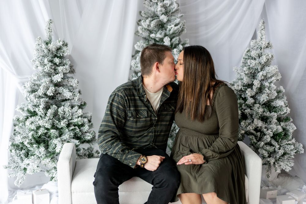 Expecting couple sharing a kiss on white bench with snow-covered Christmas trees in holiday maternity portrait.