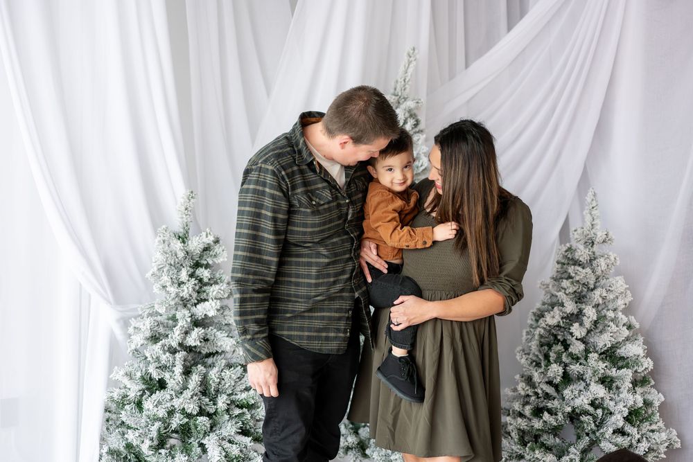 A family shares a tender moment with their young child between snow-covered Christmas trees indoors.