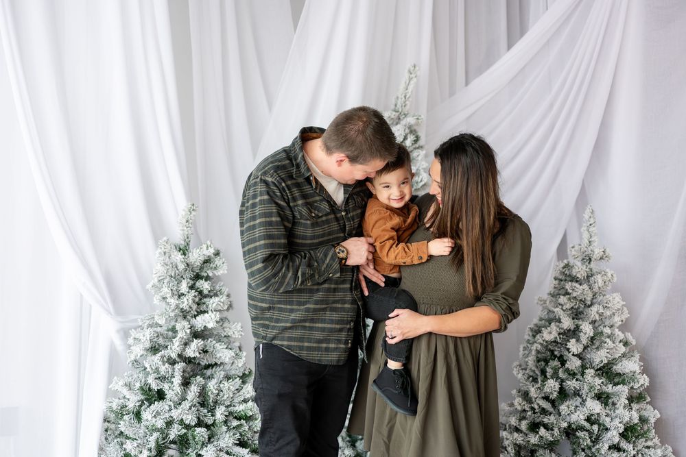Parents kiss their young son on the cheeks in a warm holiday portrait with snowy Christmas trees.