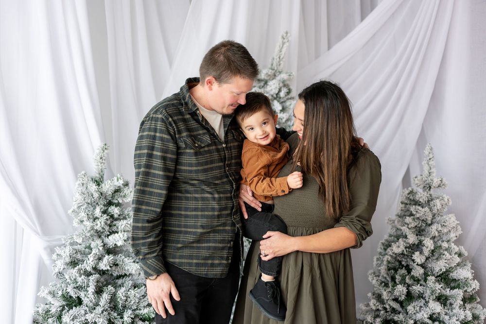 A couple lovingly gazes at their toddler son amid snow-flocked Christmas trees in a holiday portrait.