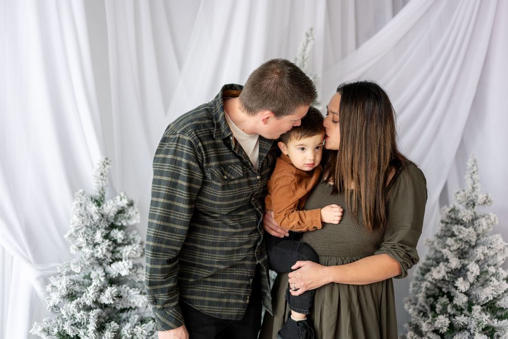 A couple shares a loving moment kissing their young child between elegant snow-flocked Christmas trees.