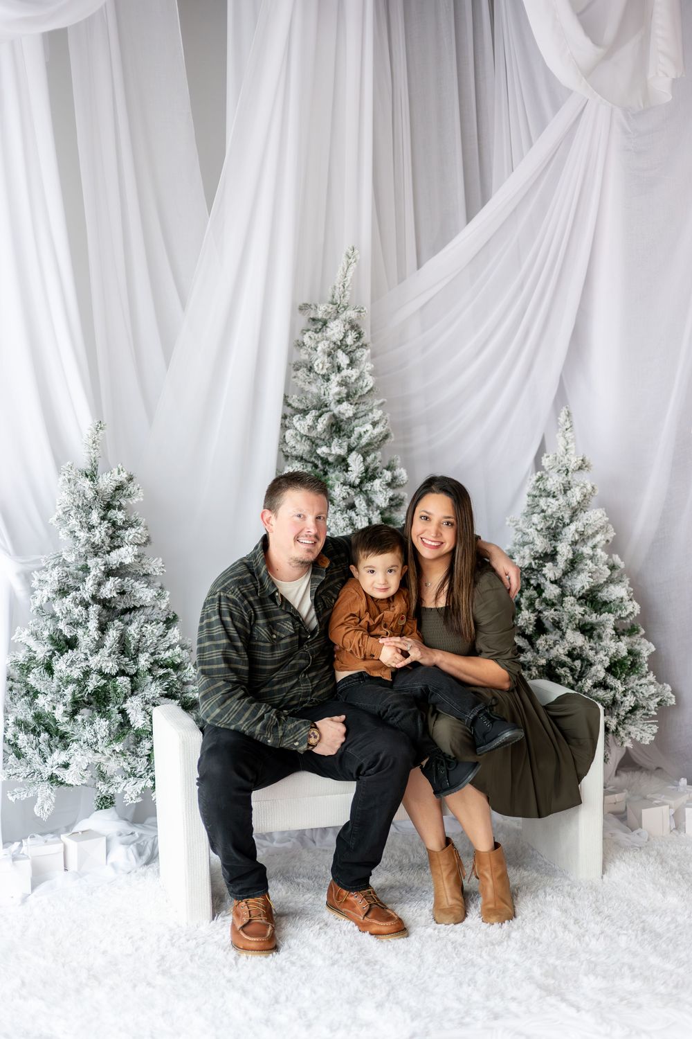 Family of three posing together on white bench surrounded by snow-flocked Christmas trees in festive studio setting.
