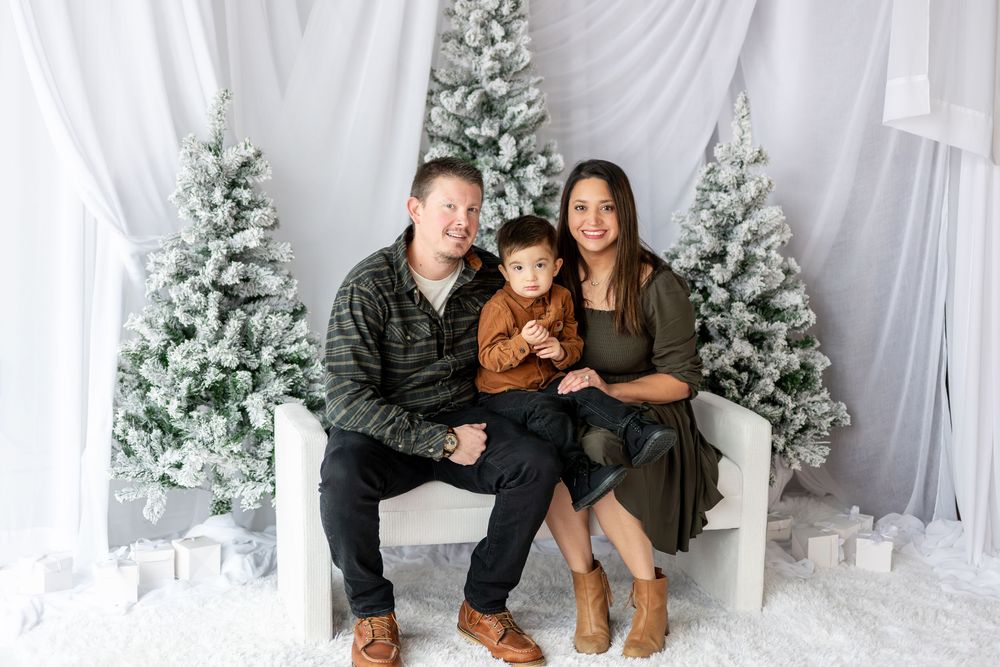A smiling family of three sits together on a white bench surrounded by snow-flocked Christmas trees.