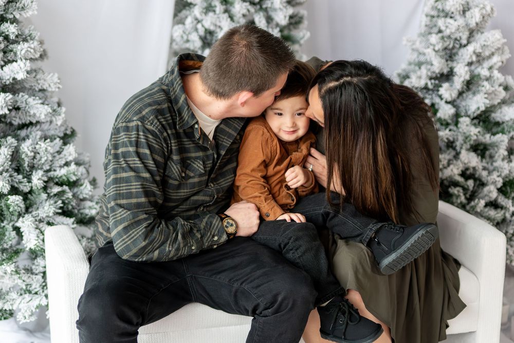 Two parents kiss their toddler on the cheeks in a sweet holiday portrait with snowy Christmas trees.