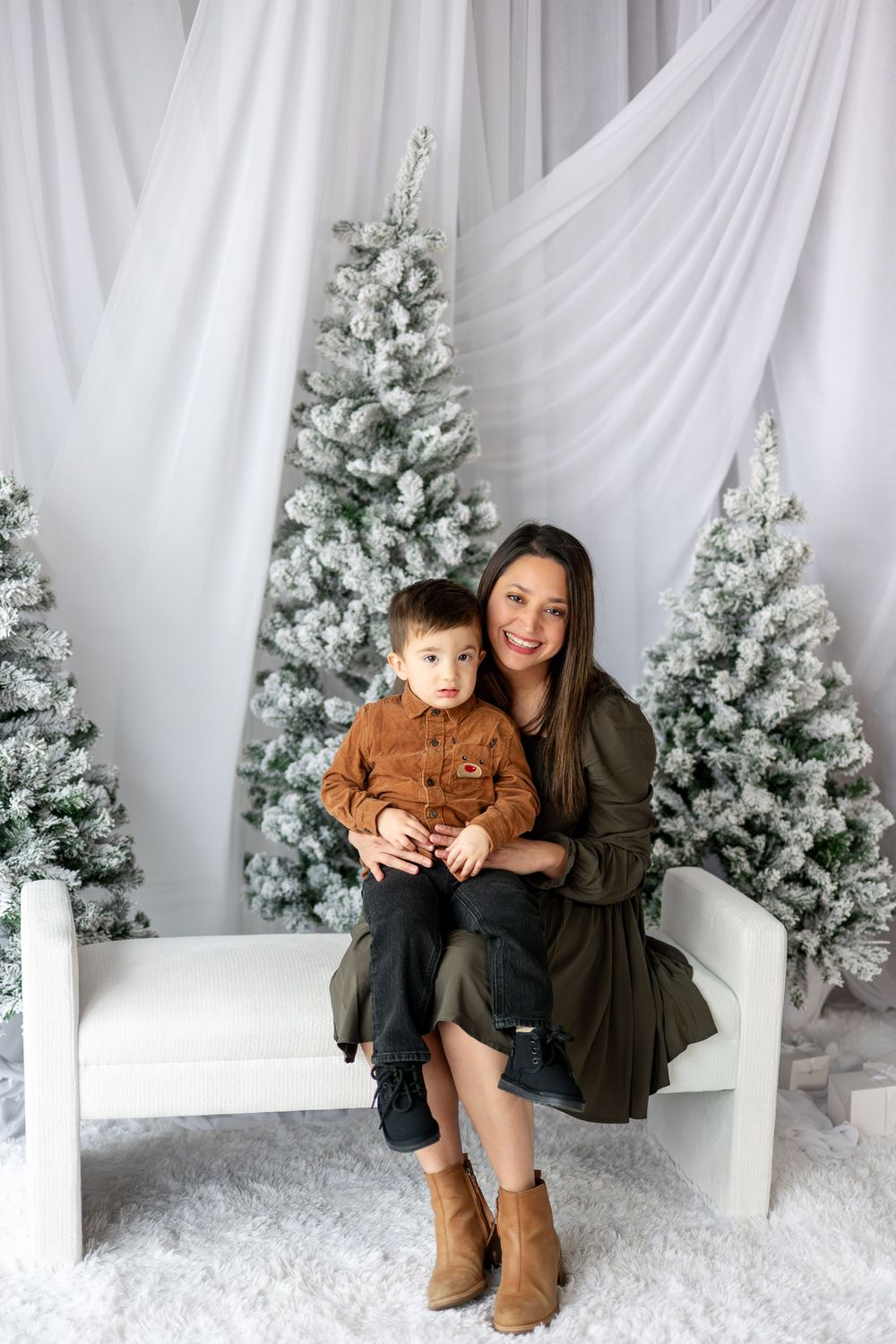 Mother and young toddler son sitting together on white bench with snow-flocked Christmas trees in studio backdrop.