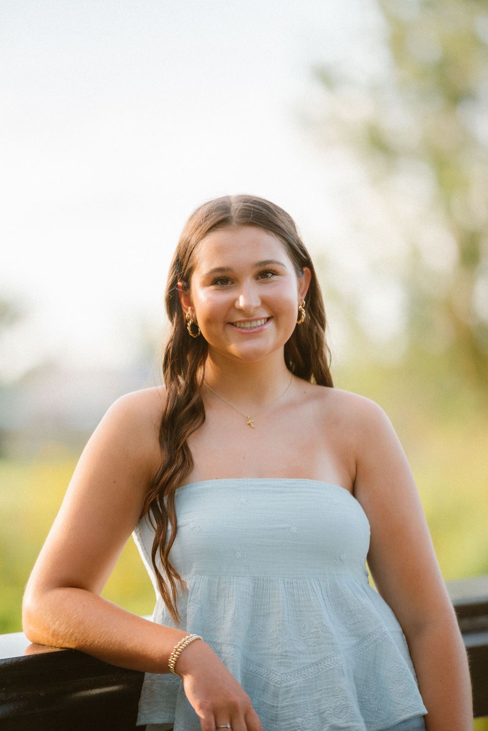 A woman in a light blue strapless top poses outdoors during golden hour with a warm natural smile.