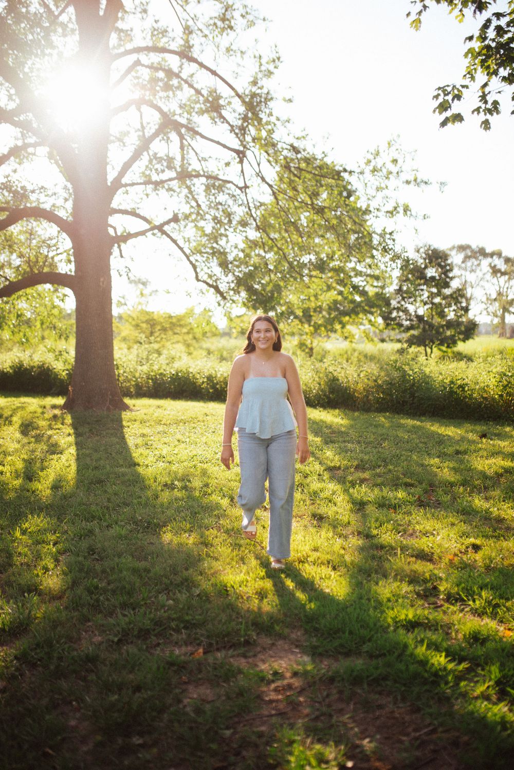 A person walks up a grassy hillside path at sunset with sunlight streaming through trees in a dreamy summer scene.