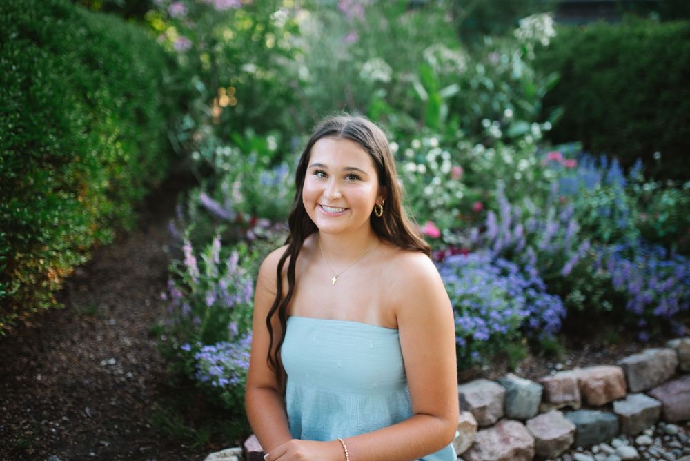 A person in a light blue dress poses in a beautiful garden with purple flowers and stone edging along the path.