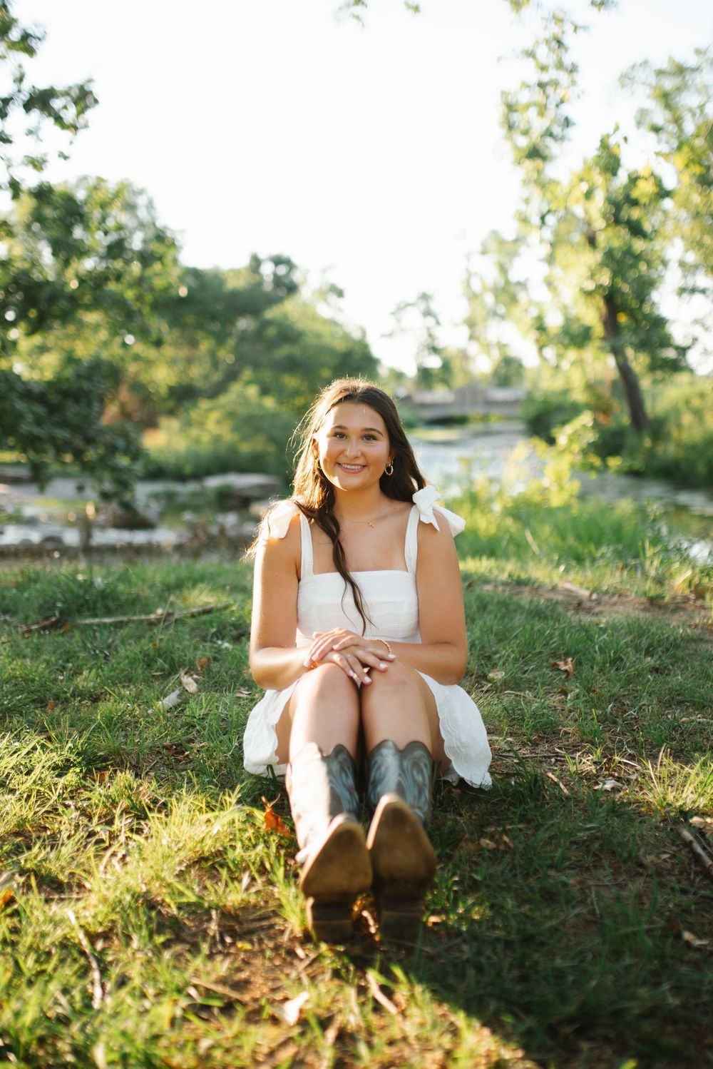 Person sitting outdoors on grassy hillside during golden hour sunlight in white summer dress.