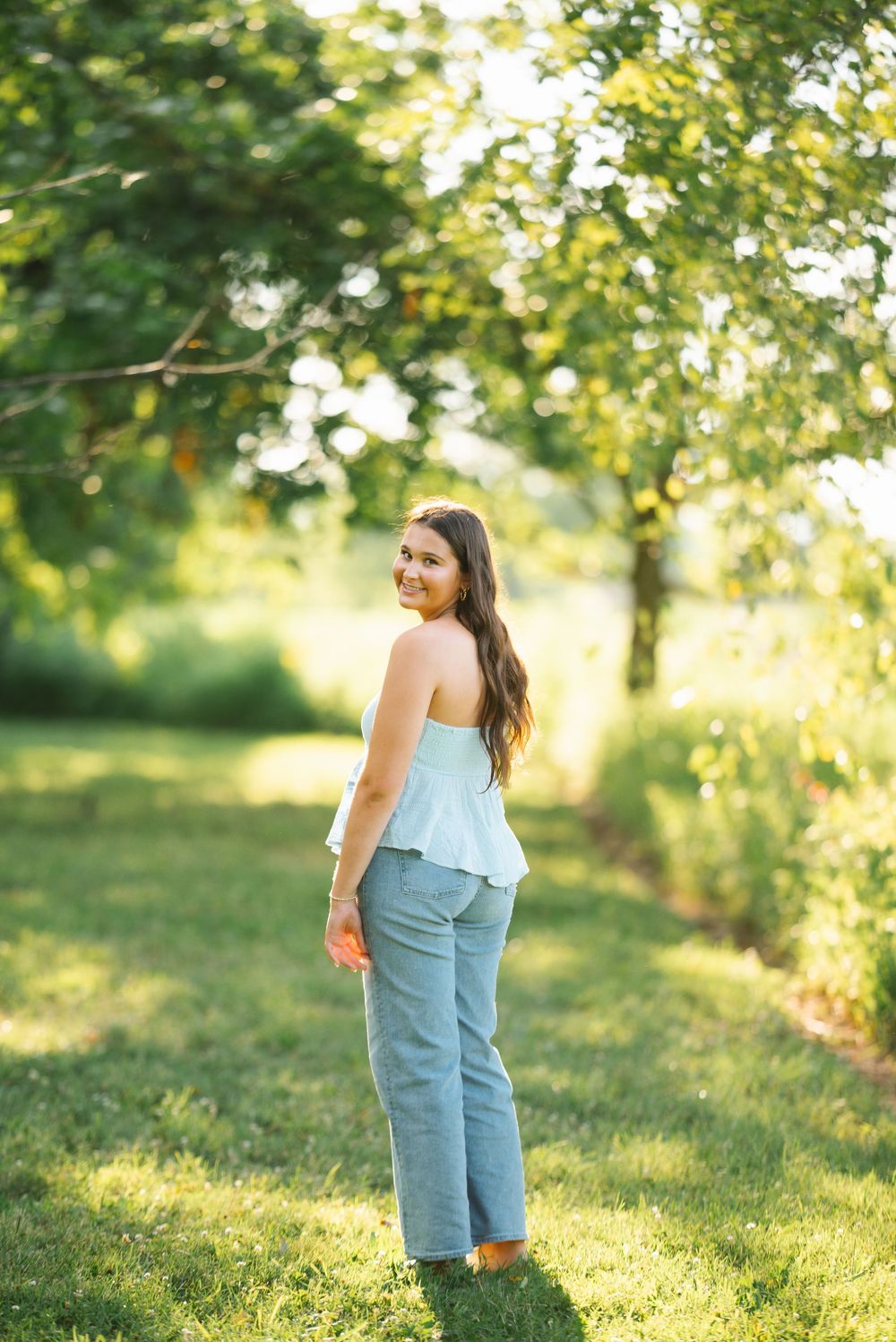 A woman in a white top and jeans walks through a sun-dappled orchard during golden hour.