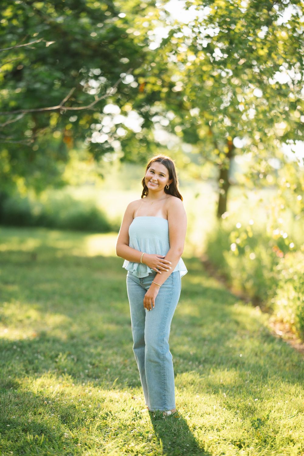 A person in a light blue top and jeans poses in a sunlit garden with green trees and grass in the background.