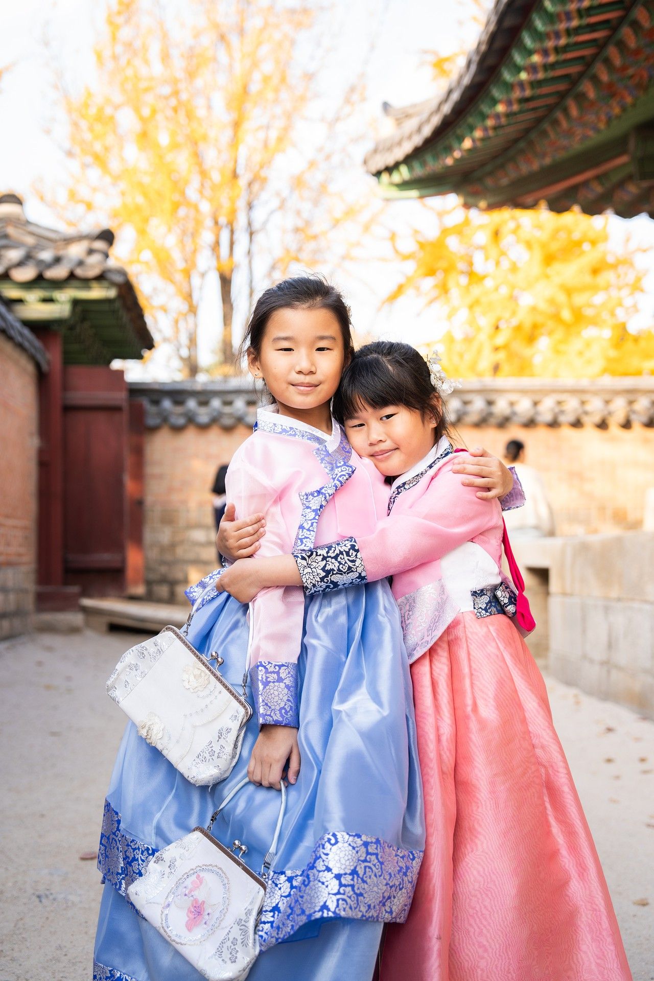 Two people in traditional Korean hanbok dresses embrace while standing in a courtyard with autumn trees.