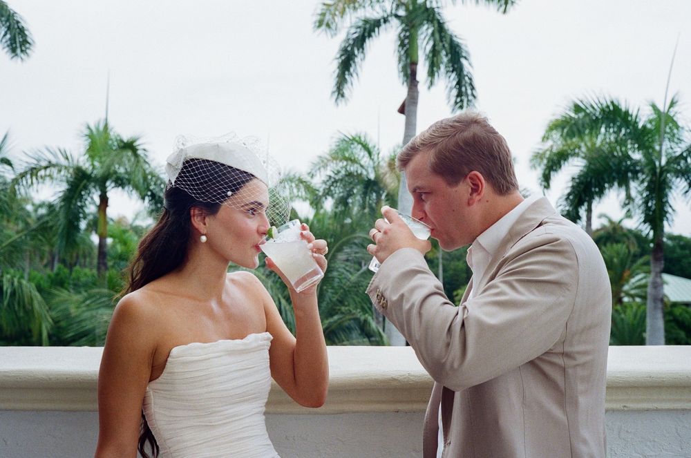 A couple in wedding attire toasts with champagne glasses in a tropical garden setting.
