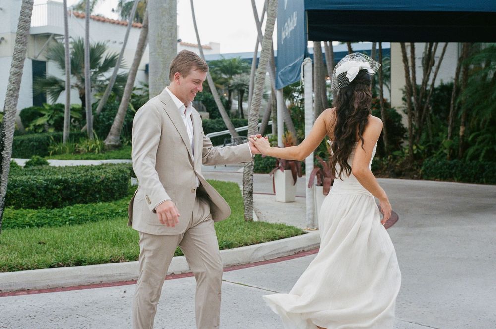 A wedding couple walks down a palm-lined path in tropical beige and white attire on their special day.
