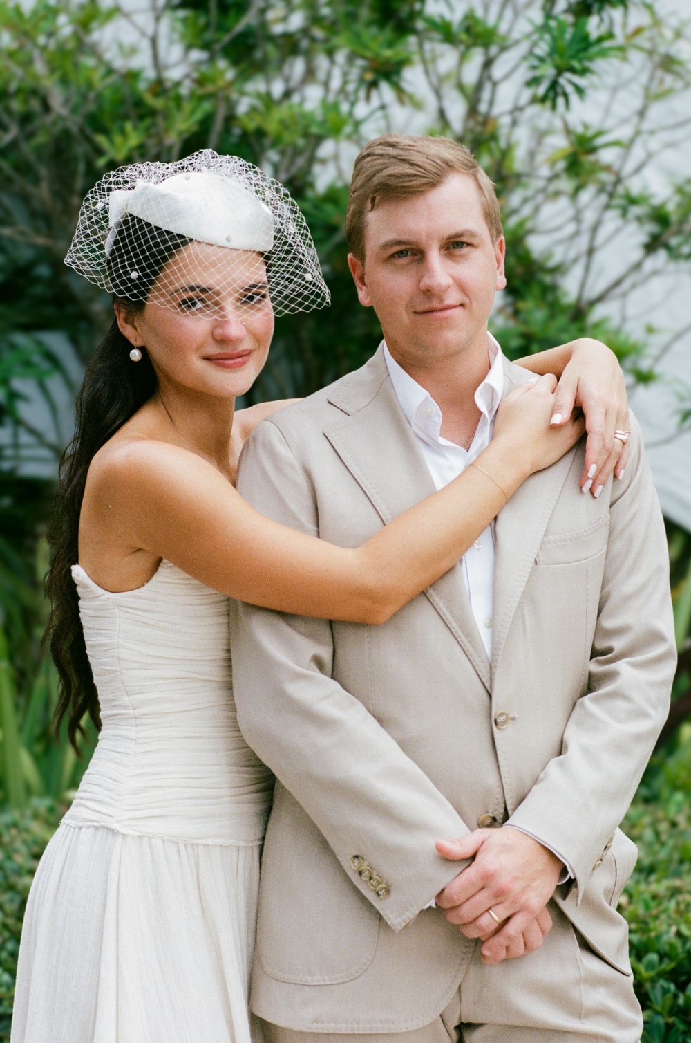 Wedding couple poses together outdoors, bride in white dress and groom in light beige suit against greenery.