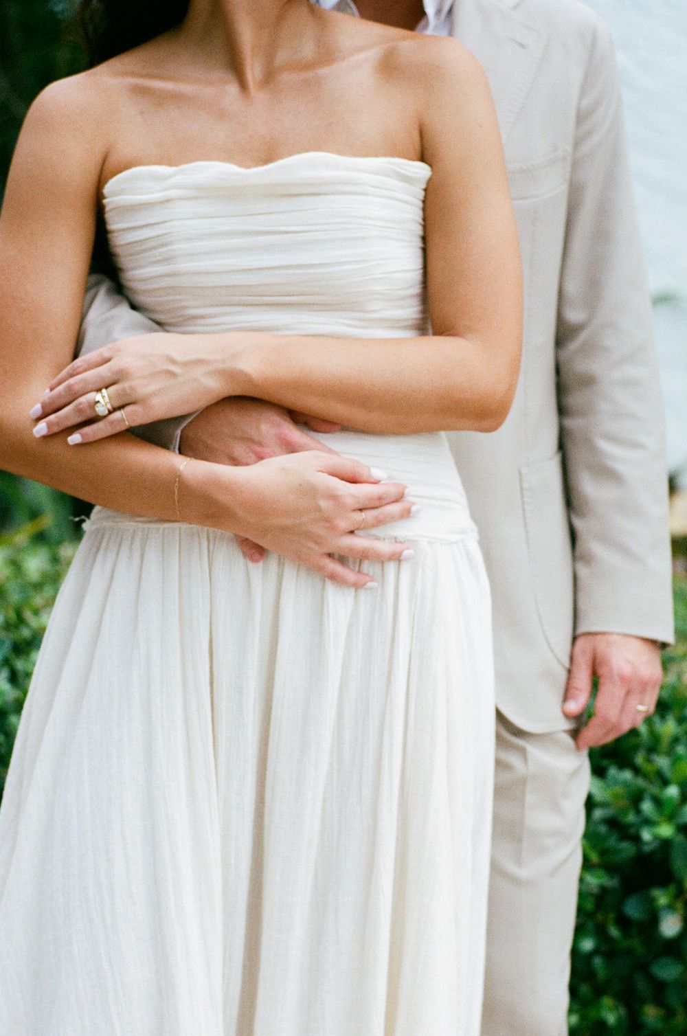 Close-up detail shots of a bride in a white strapless wedding dress showing hands and torso against green foliage.