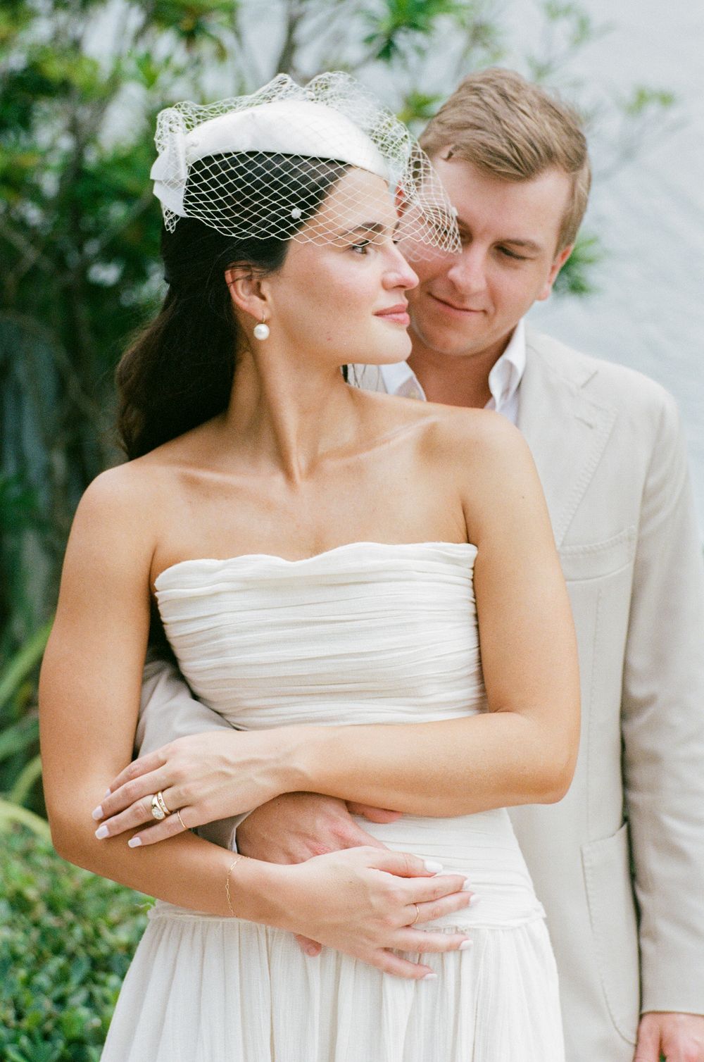 A romantic wedding couple embraces outdoors, with the bride in a strapless white dress and birdcage veil.