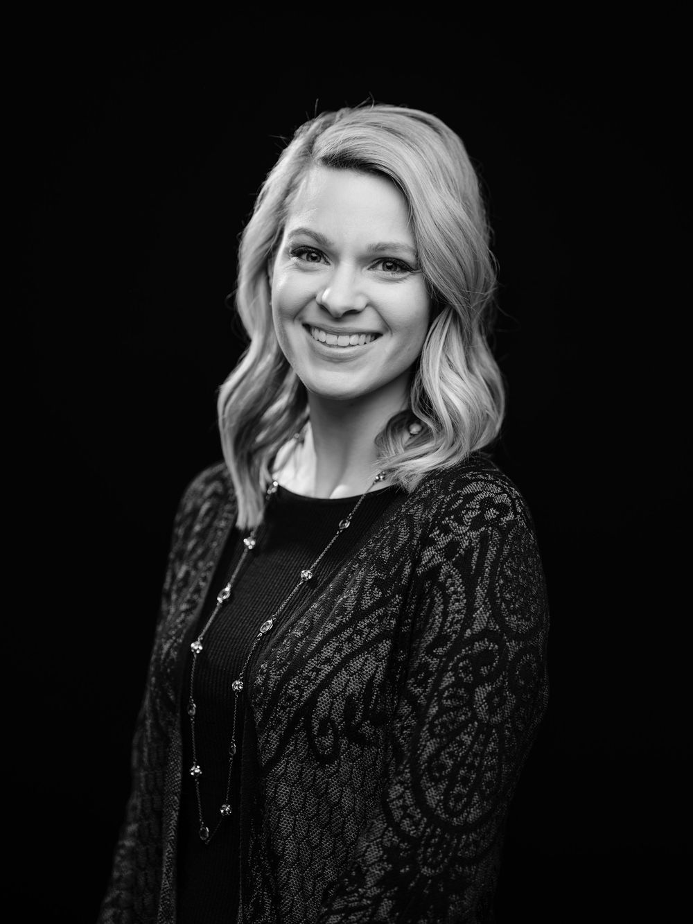 Professional studio portrait against black background showing elegant black lace jacket and warm smile.