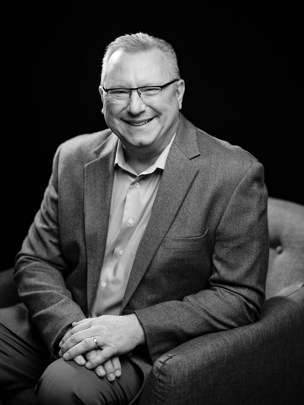 Professional black and white portrait of a person in glasses and blazer smiling warmly against dark background.