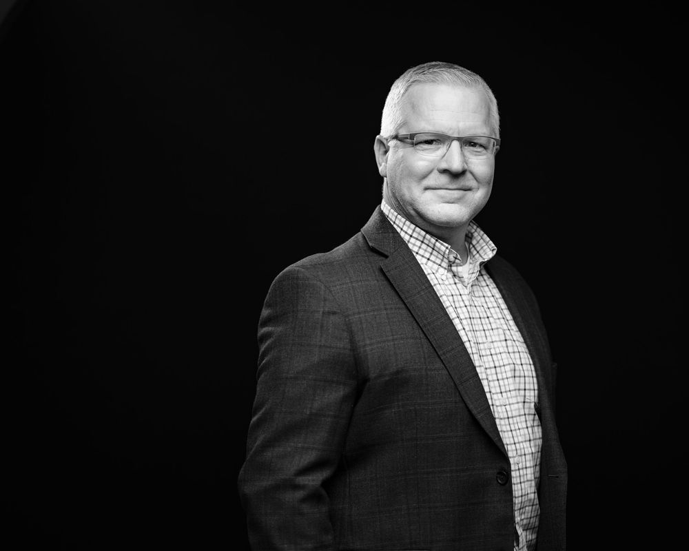 Professional headshot portrait of person in dark suit and checkered shirt against black background.