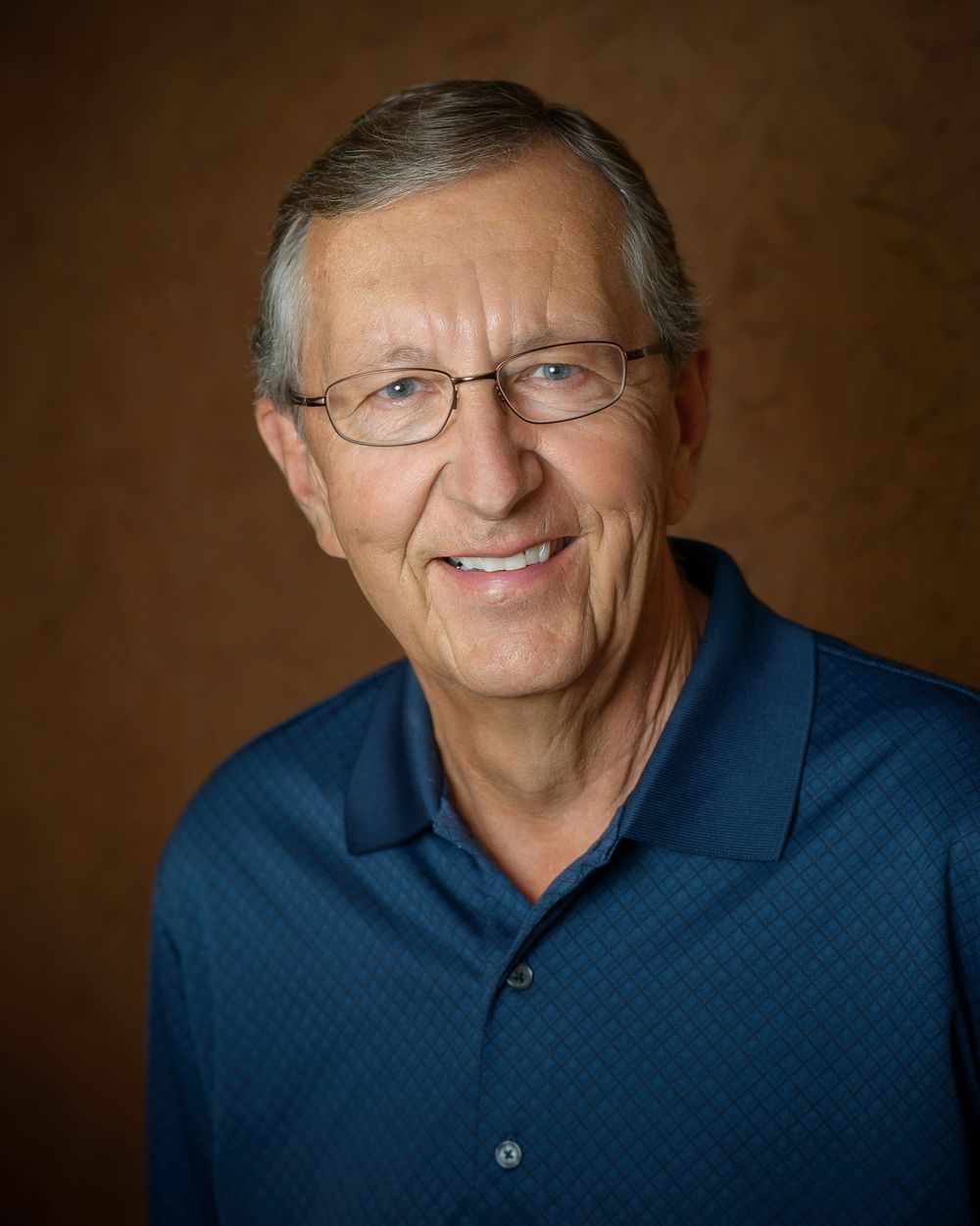 Professional headshot portraits of someone wearing glasses and a navy polo shirt against a brown background.