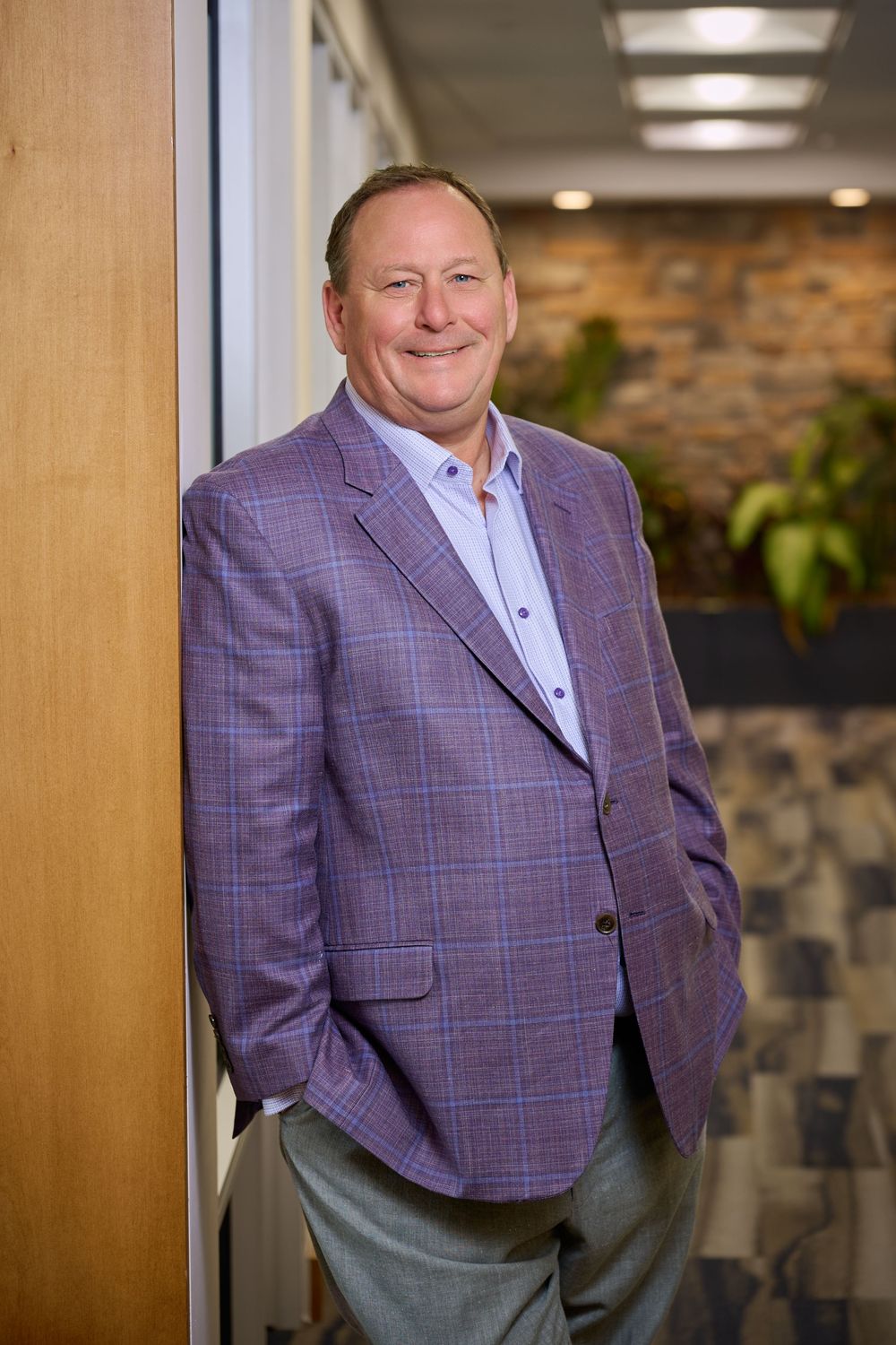 Professional portrait of someone in a purple plaid blazer and light blue dress shirt standing in an office hallway.
